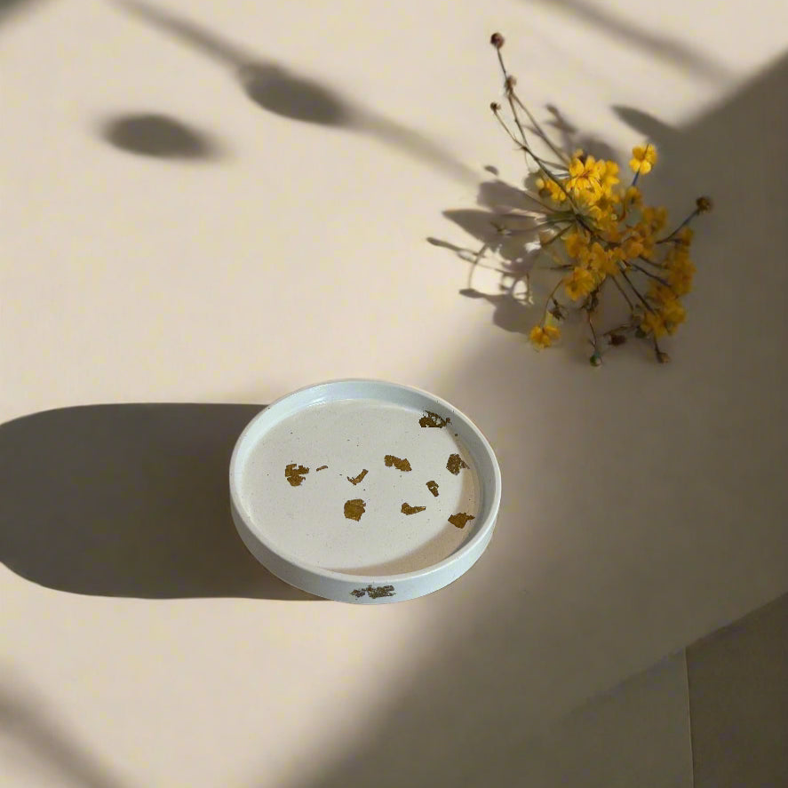A white, round, porcelain decorative dish with gold accents and floral patterns, displayed on a table with a shadow of a plant over it.