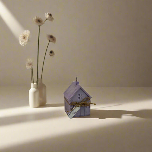 A small, handmade candle shaped like a house with a matches stick on top, placed on a white surface, with a white vase containing flowers in the background.