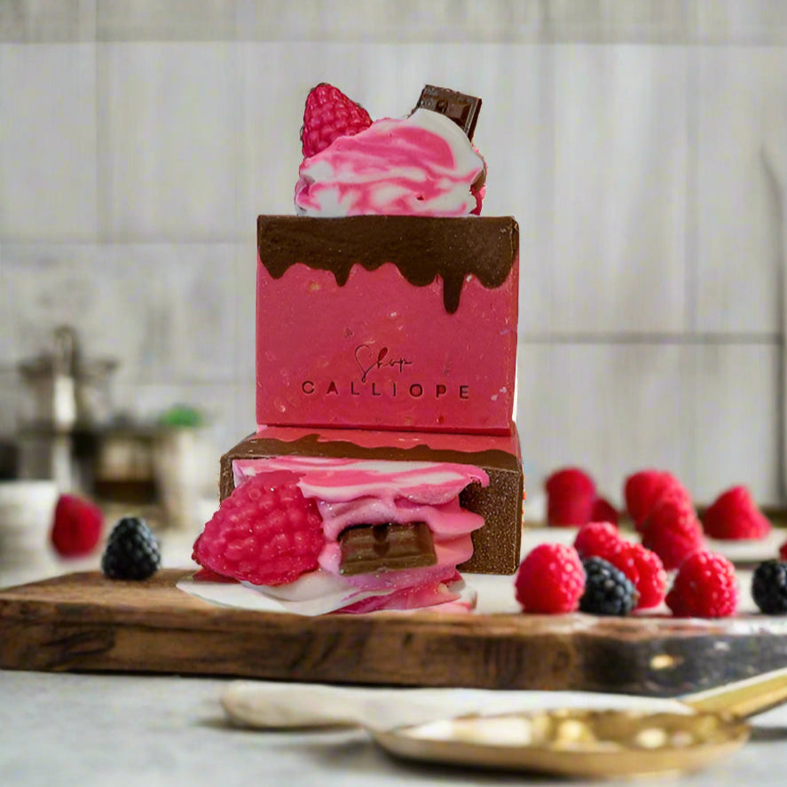 Handmade soap bar with a pink base, decorated with chocolate accents and fresh raspberries, placed on a wooden board with more raspberries and a chocolate bar in the background.