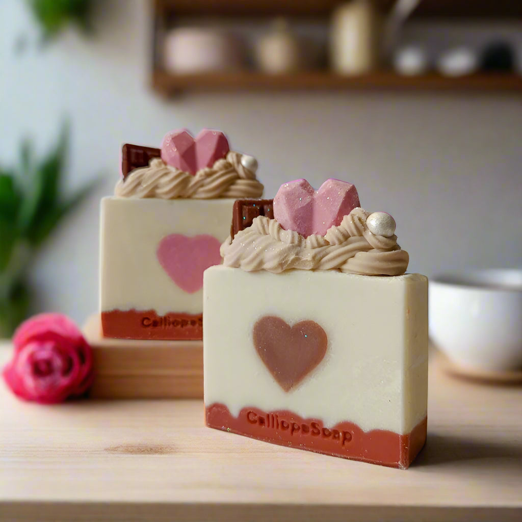 Handmade soap bars with heart shapes and decorative toppings, displayed on a wooden surface with a rose and kitchen backdrop.
