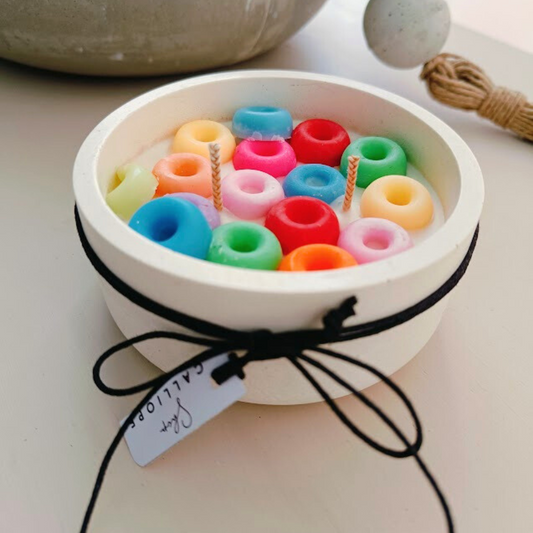 A colorful soy wax candle shaped like a bowl of cereal loops, set in a white concrete container, with a black lid tied with a string.