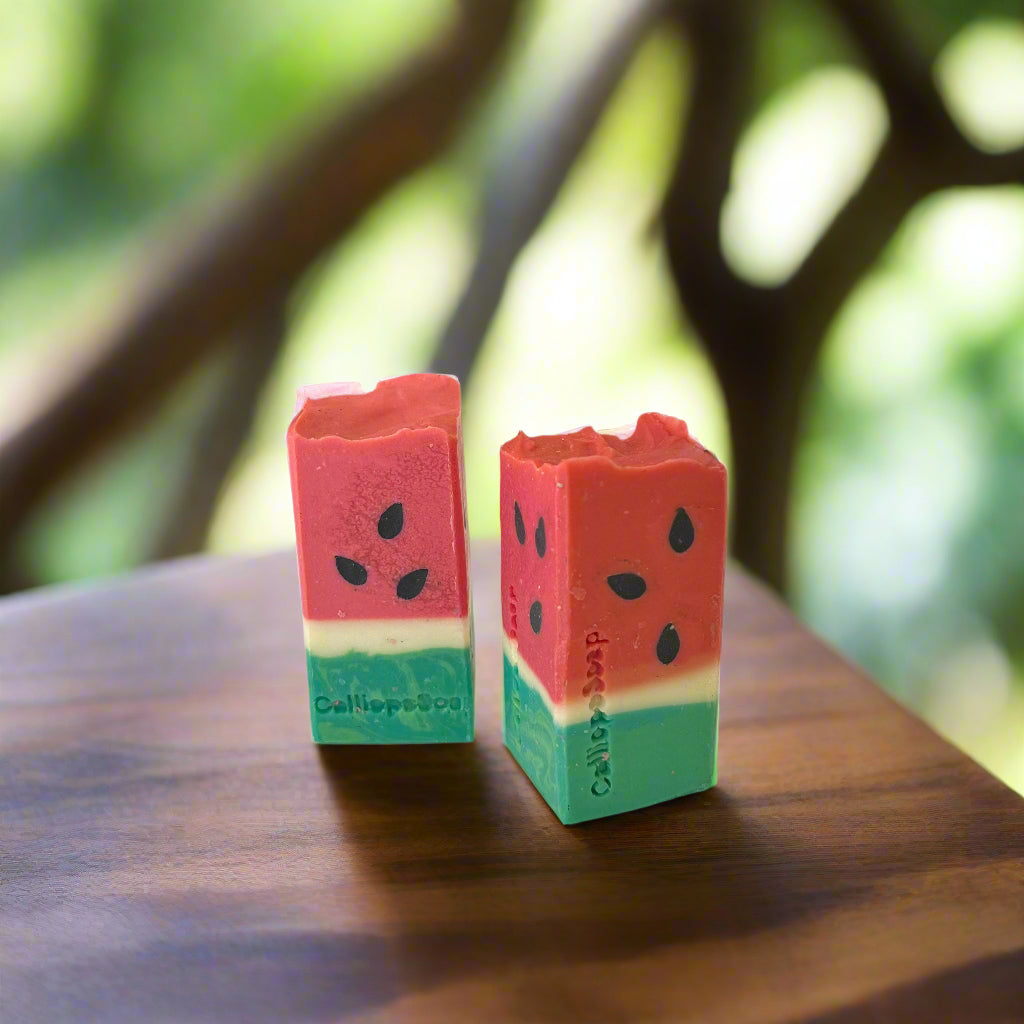 Two watermelon-themed soap bars on a wooden surface with a blurred natural background