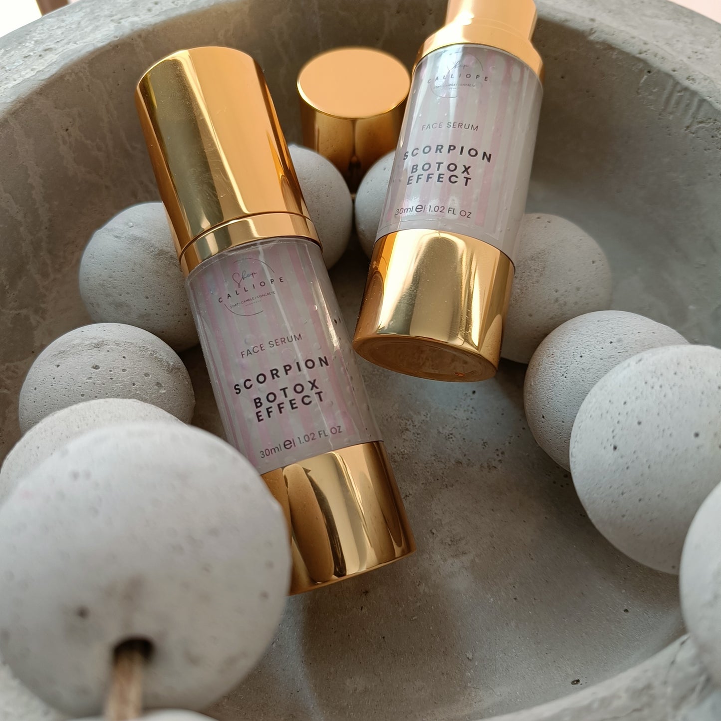 A gold-colored bottle with a pump dispenser labeled 'Scorpion Botox Effect Serum' in a cement bowl with cement beads.