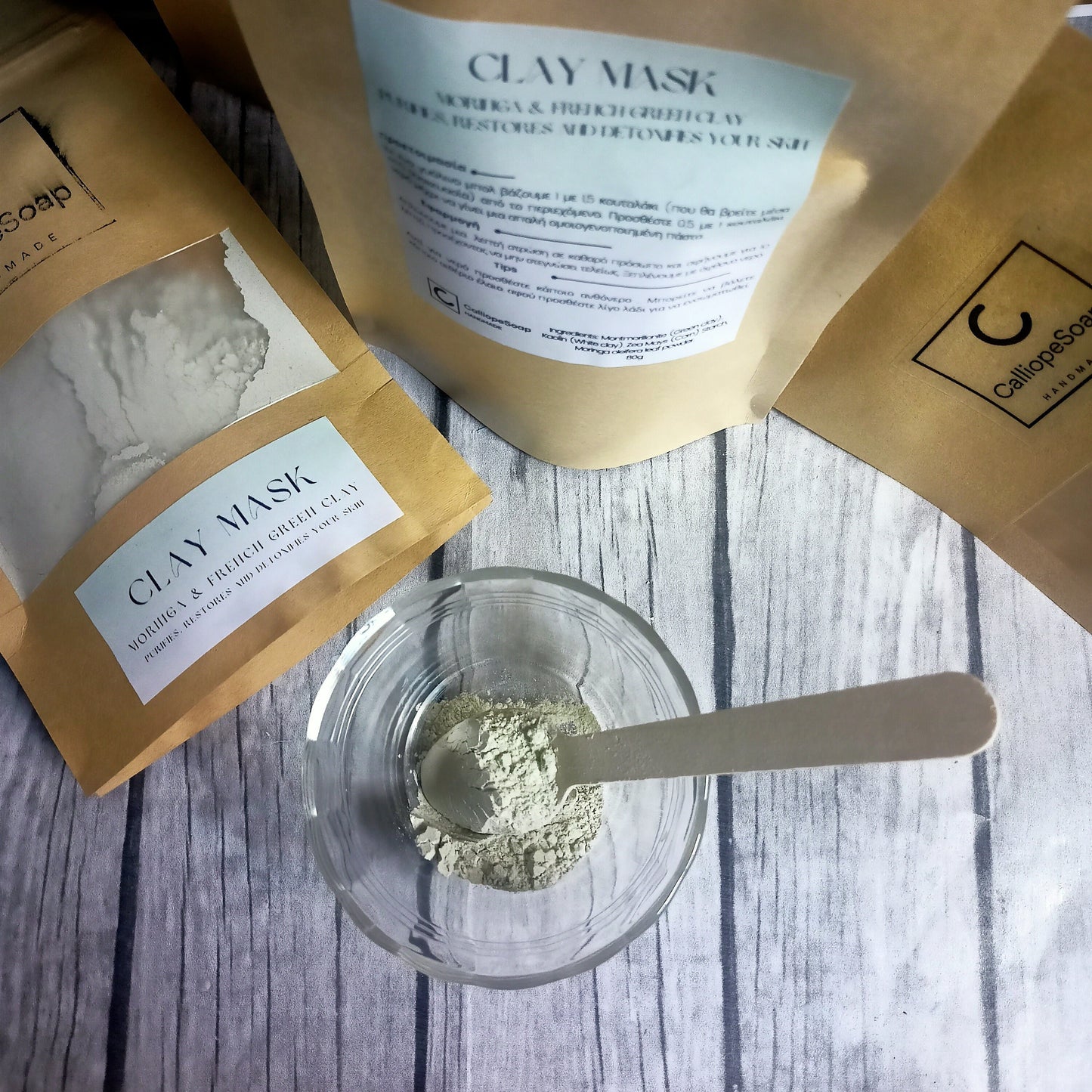 The image shows packets of clay mask powder with a small scoop and a glass bowl on a wooden surface, indicating a spa or skincare product.