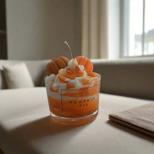 A clear glass candle with orange and white wax layers, labeled 'Pumpkin Pie', placed on a surface with a blurred background.