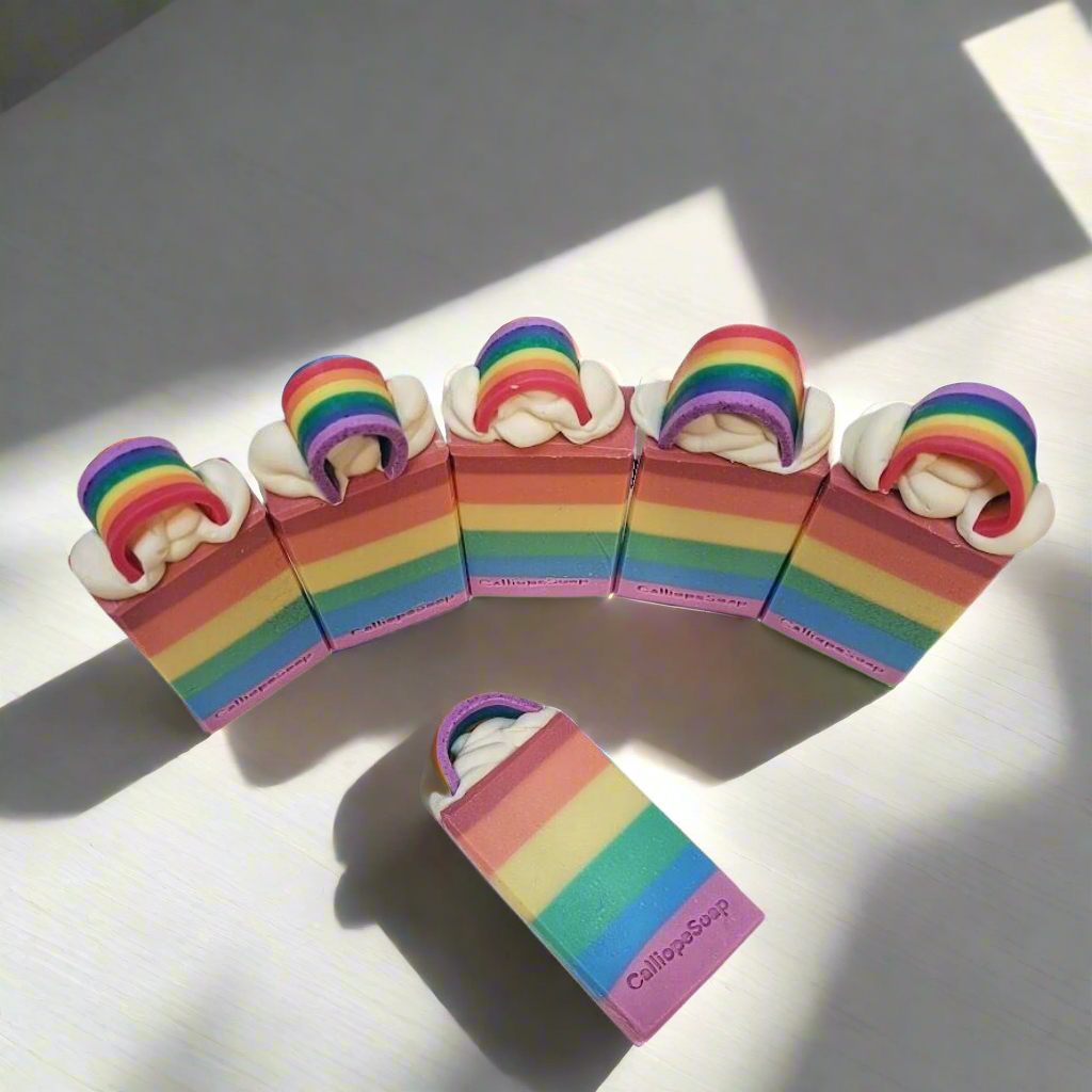 A series of rainbow-colored bar soaps with white frosting on top, displayed on a surface with a shadow falling over part of the image.