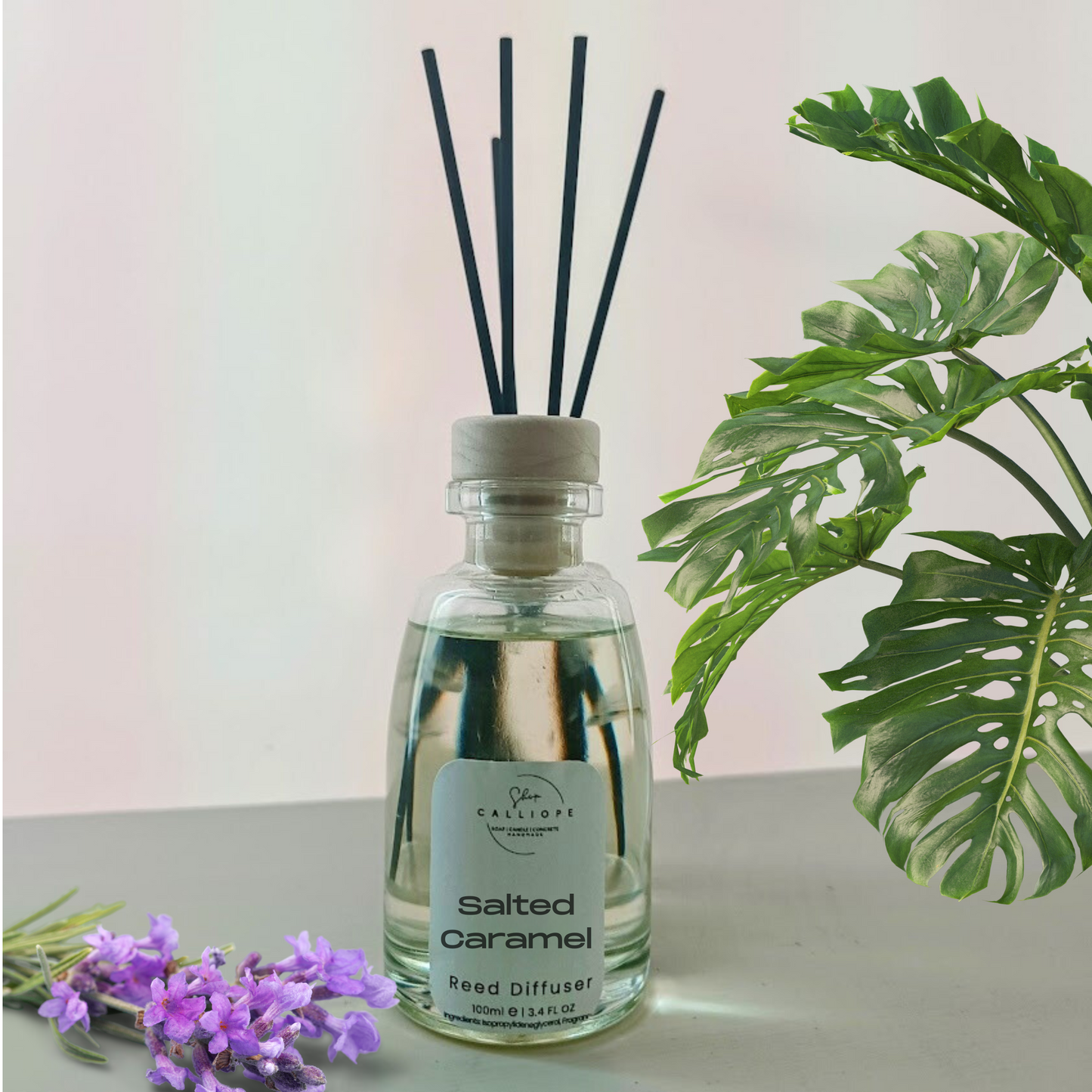 Glass diffuser bottle labeled 'Salted Caramel' with black sticks on a light surface with green leaves and purple flowers in the background.