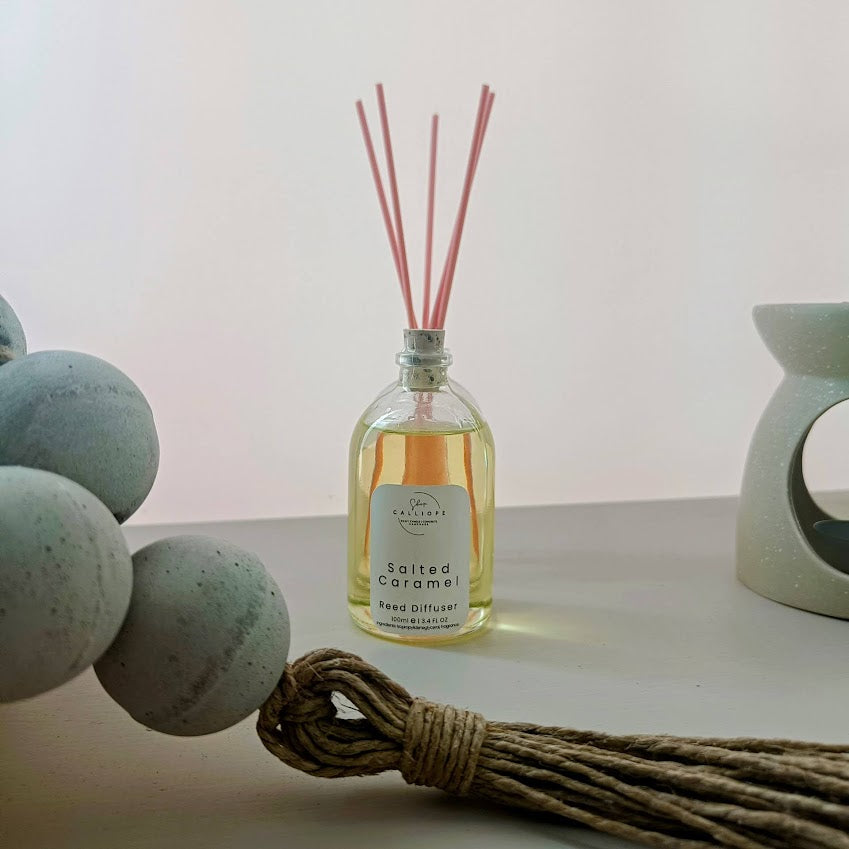 A clear glass reed diffuser bottle filled with fragrance oil, positioned on a table with a few reeds in different colors, a blue stone coaster, and a rope-like object partially visible in the foreground.