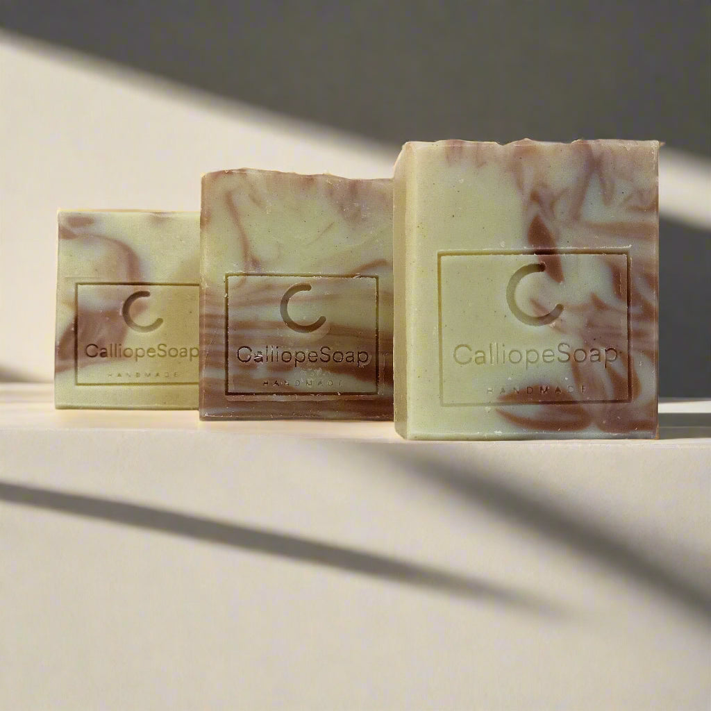 Three bars of handcrafted soap with a marbled pattern in shades of beige and white, each with a label indicating the brand 'Calliope Soap'.