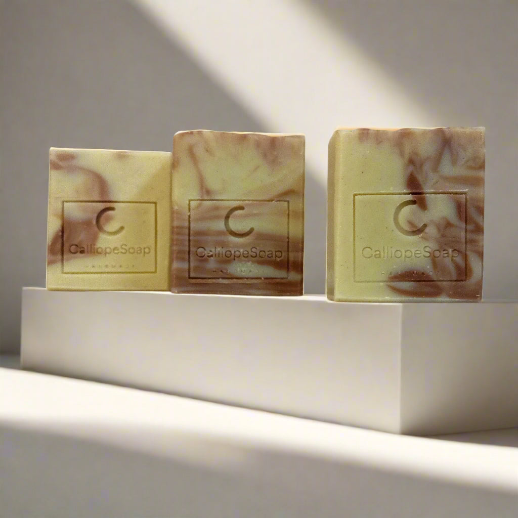 Three bars of handcrafted soap with a marbled pattern in shades of beige and white, each with a label indicating the brand 'Calliope Soap'.