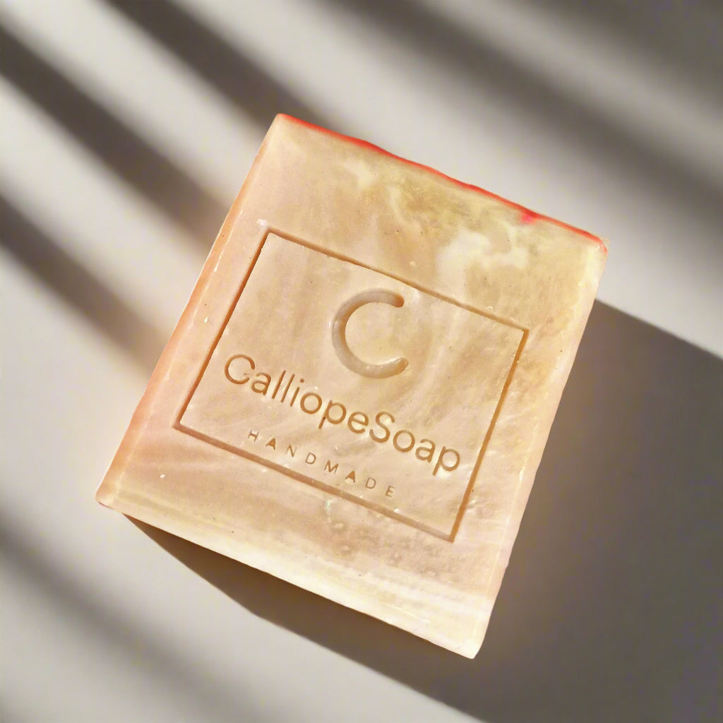 A bar of handmade soap with red clay, branded with 'calliopeshop.com', placed on a wooden surface with a plant in the background.