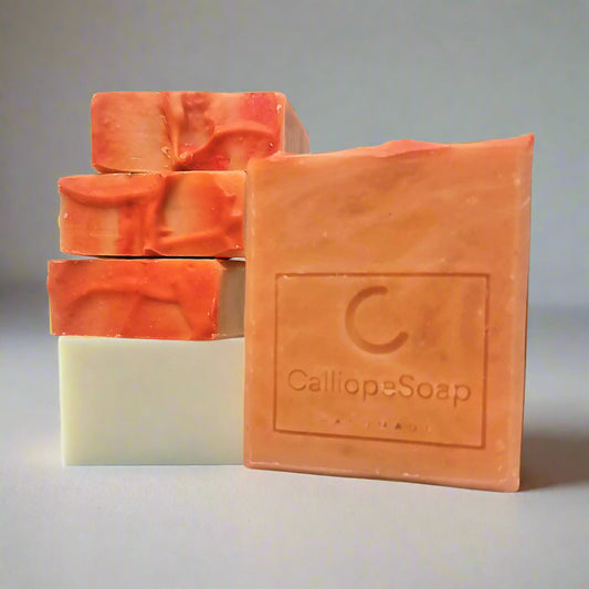 A bar of handmade soap with red clay, branded with 'calliopeshop.com', placed on a wooden surface with a plant in the background.