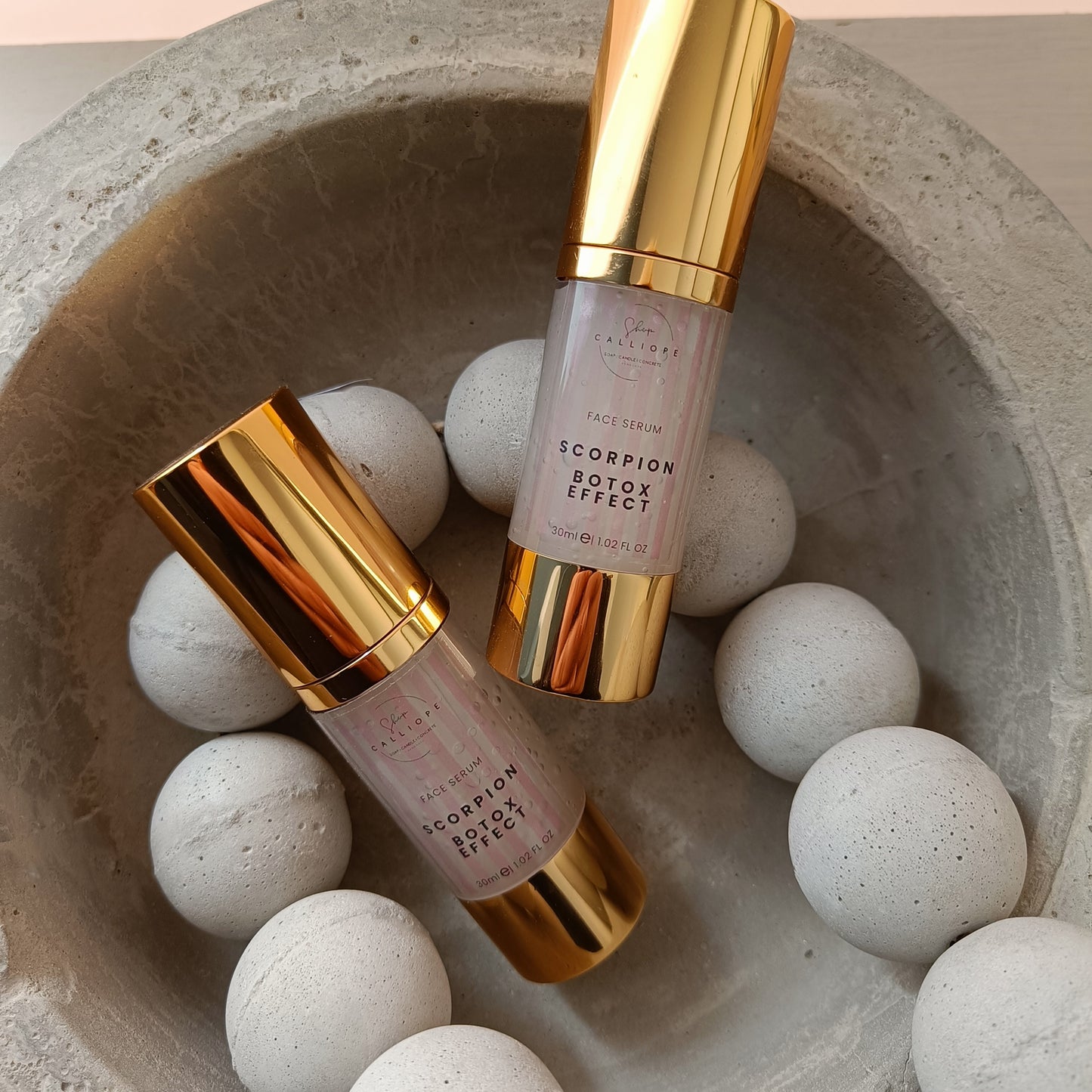 A gold-colored bottle with a pump dispenser labeled 'Scorpion Botox Effect Serum' in a cement bowl with cement beads.
