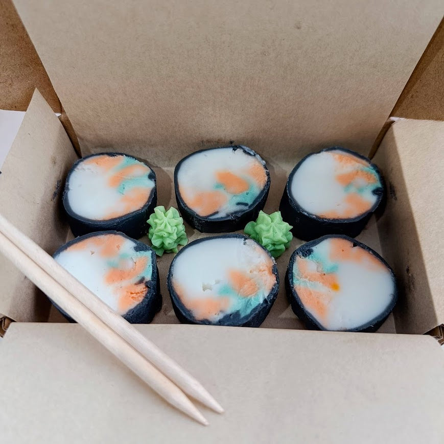 Sushi-shaped candles in a cardboard box with chopsticks on a beige surface