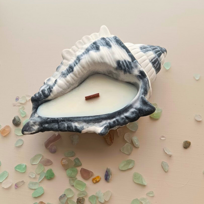 A handpoured soy candle with a swirled pattern of dark and light grey, resembling a seashell, on a light background with sea shells.