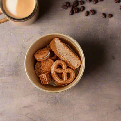 A cup filled with wax melts shaped like various baking items such as cookies and pretzels, placed on a grey surface with coffee beans scattered around.