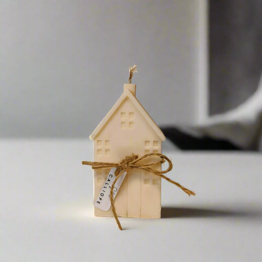 Three handmade soy wax candles designed to look like small houses, in beige, purple, and green colors, displayed on a white platform with twine decorations.