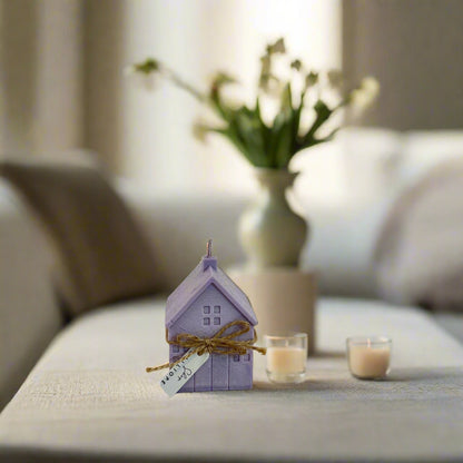 A purple candle shaped like a small house with a bow on top, placed on a table in a cozy home setting, with a vase of flowers and a lit candle in the background.