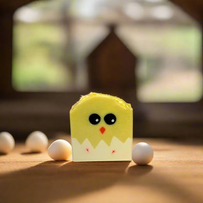 Handmade yellow soap bar with black eyes and a smiling face resembling a chicken, placed among white eggs on a wooden surface with a window in the background.