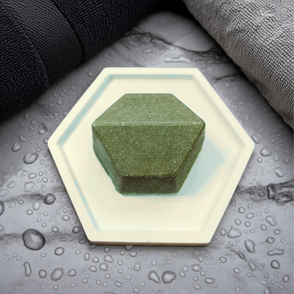 Green bar of soap on a hexagonal white soap dish with water droplets on a gray surface.