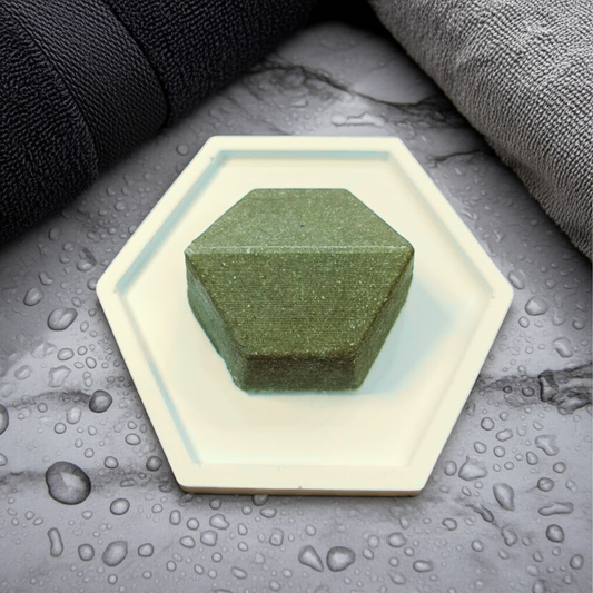 Green bar of soap on a hexagonal white soap dish with water droplets on a gray surface.
