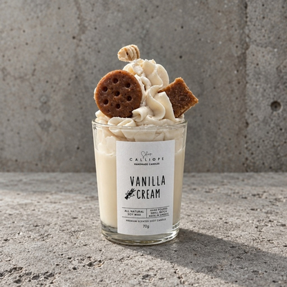 Ένα κερί σε γυάλινο βάζο με την ετικέτα 'Vanilla Cream' από την Calliope Handmade Candles, γεμιστό με κρεμώδες κερί βανίλιας και γαρνιρισμένο με σαντιγί, ένα μπισκότο και ένα γκοφρετάκι, τοποθετημένο σε μια ανάγλυφη επιφάνεια από  τσιμέντο. 