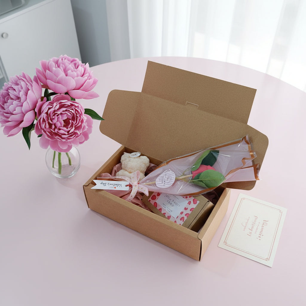 Open cardboard box with floral arrangement and card on a pink surface, pink flowers in the background.