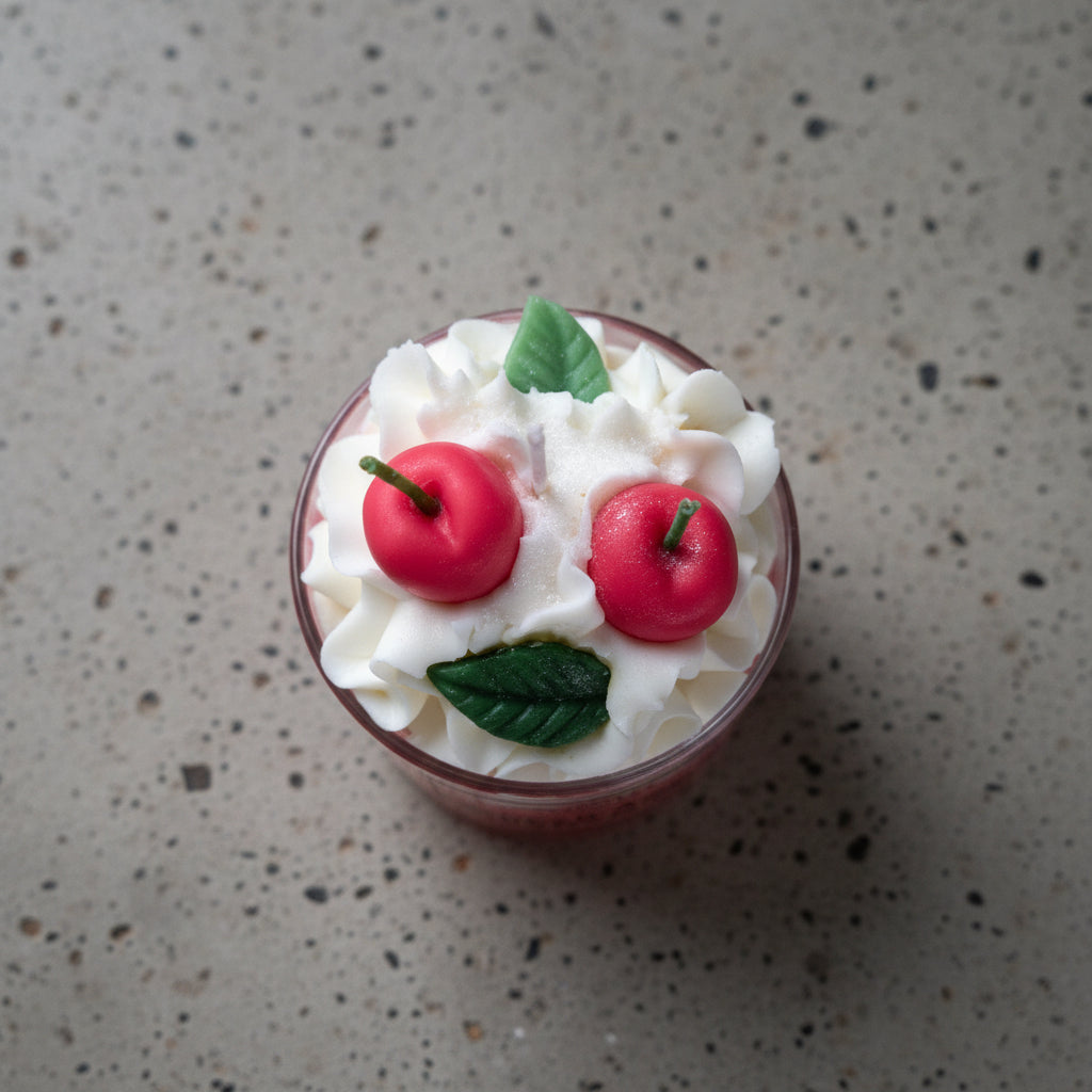 Λεπτομέρεια από το κερί σε ποτήρι spiced apple cinnamon , λευκή σαντιγί με δυο μήλα και δυο πράσινα φύλλα από κερί διακοσμητικά στην κορυφή, πάνω σε γκρι επιφάνεια