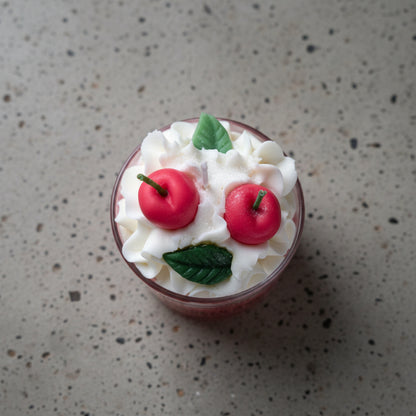 Λεπτομέρεια από το κερί σε ποτήρι spiced apple cinnamon , λευκή σαντιγί με δυο μήλα και δυο πράσινα φύλλα από κερί διακοσμητικά στην κορυφή, πάνω σε γκρι επιφάνεια