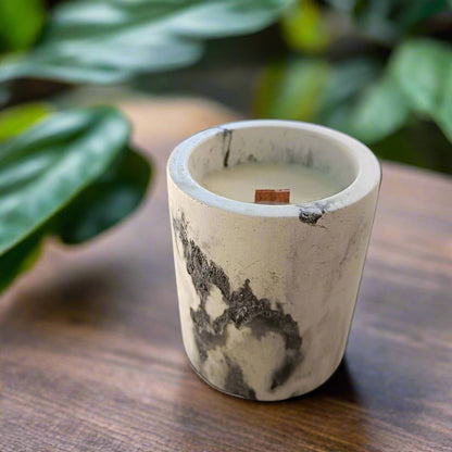 A handmade concrete candle bowl with a burning soy candle inside, set on a wooden surface with green foliage in the background.