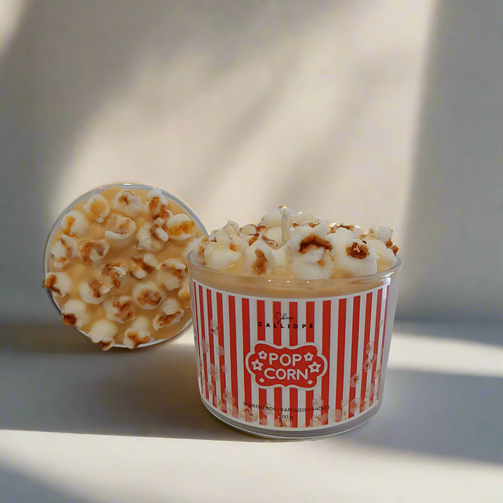 Candle in a container labeled 'Pop Corn' with popcorn design on a neutral background