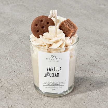Ένα χειροποίητο κερί με άρωμα vanilla cream από την Calliope σε διάφανο γυάλινο βάζο, γαρνιρισμένο με σαντιγί, ένα μπισκότο γεμιστό και ένα κομμάτι βάφλας, τοποθετημένο σε ένα ανάγλυφο γκρι φόντο.