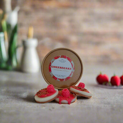 A set of handmade wax melts designed to resemble cheesecake, with a label indicating the scent. The melts are displayed on a neutral background alongside a ceramic diffuser and fresh raspberries.