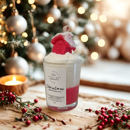 Christmas-themed candle with snowy red house design on top, surrounded by festive decorations.