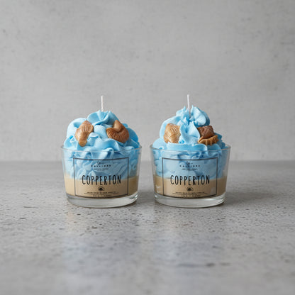 Two blue-themed candles with shell figures in glass containers on a concrete surface.
