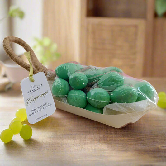 Green grape-shaped soaps in a clear plastic bag with a wooden basket on a light surface, with pink bottles in the background.
