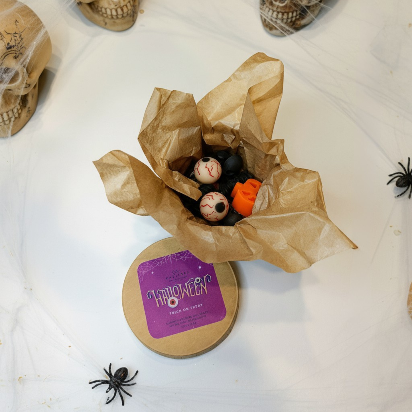 Halloween-themed wax melts in craft box with small pumpkins, bat, eyeballs  and a skull on a white surface.