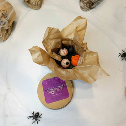 Halloween-themed wax melts in craft box with small pumpkins, bat, eyeballs  and a skull on a white surface.
