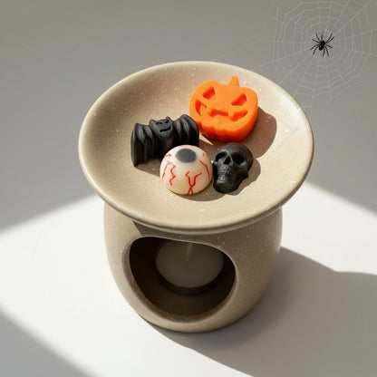 Halloween wax melts on a ceramic wax burner, including a jack-o'-lantern, bat, eyeball, and skull.