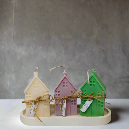 Three handmade soy wax candles designed to look like small houses, in beige, purple, and green colors, displayed on a white platform with twine decorations.