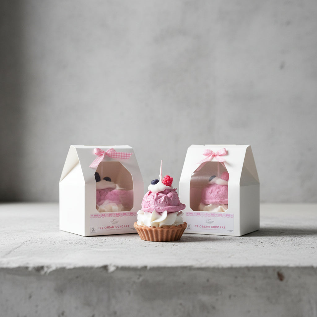 Cupcake candle with pink frosting and white cream, displayed in front of its packaging on a concret surface.