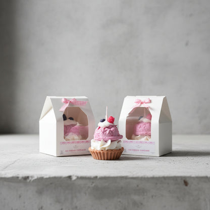 Cupcake candle with pink frosting and white cream, displayed in front of its packaging on a concret surface.