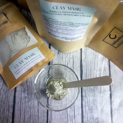 The image shows packets of clay mask powder with a small scoop and a glass bowl on a wooden surface, indicating a spa or skincare product.