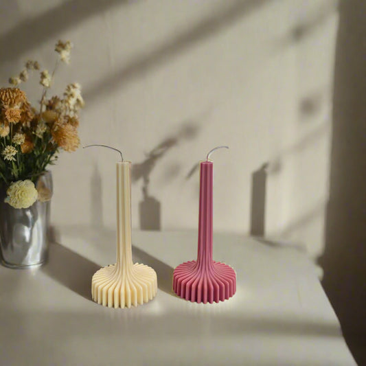 Two ribbed pillar candles, one beige and one pink, placed on a surface with a vase of dried flowers in the background.