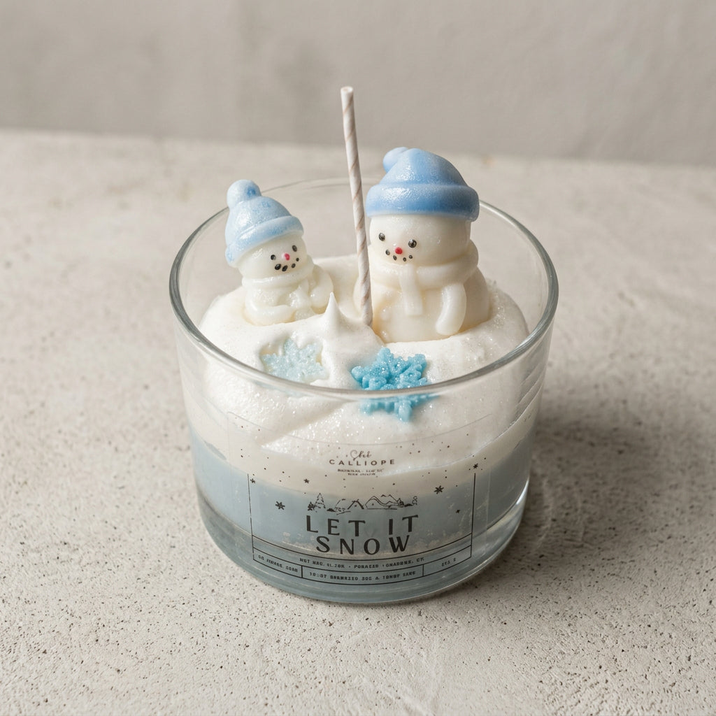 A glass jar candle with a white and light blue gradient base, featuring two snowman figurines wearing blue hats on top. The front label reads 'LET IT SNOW' and shows the brand'.
