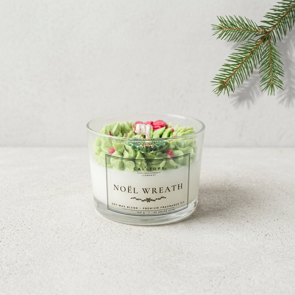 A glass jar candle labeled 'NOÉL WREATH' sits on a light stone surface against a pale wall, with green pine branches in the upper right corner. The candle has white wax and green textured decorative elements on top, with small pink and red accents.