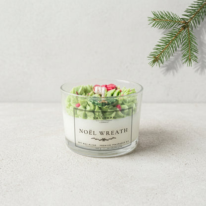 A glass jar candle labeled 'NOÉL WREATH' sits on a light stone surface against a pale wall, with green pine branches in the upper right corner. The candle has white wax and green textured decorative elements on top, with small pink and red accents.