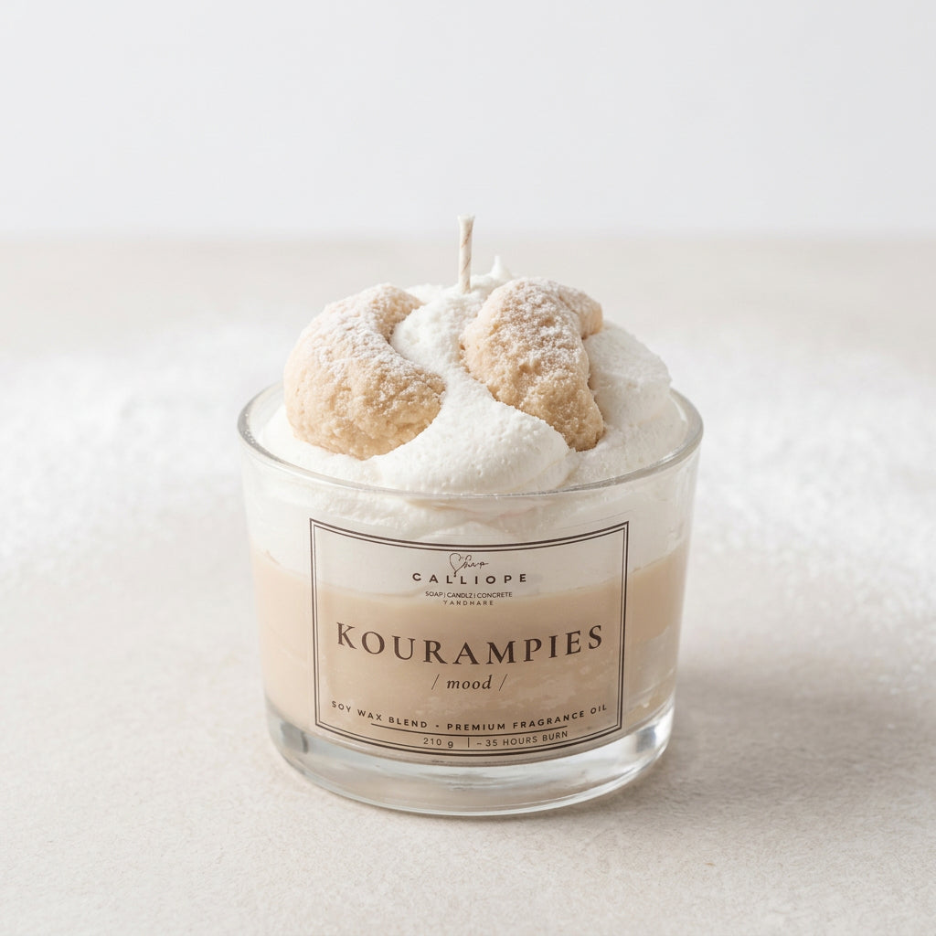 A glass jar candle labeled 'Galliope Kourampies / mood' filled with white wax and topped with two sugar-dusted koyrampiedes, with a single wick visible against a light gray background.