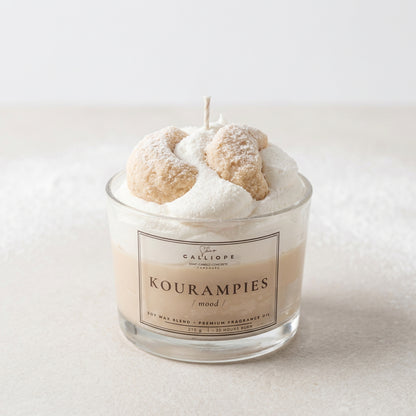 A glass jar candle labeled 'Galliope Kourampies / mood' filled with white wax and topped with two sugar-dusted koyrampiedes, with a single wick visible against a light gray background.