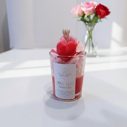 Candle with heart and a 'Love shot, CalliopeShop' label on a white surface, with a vase of flowers in the background.